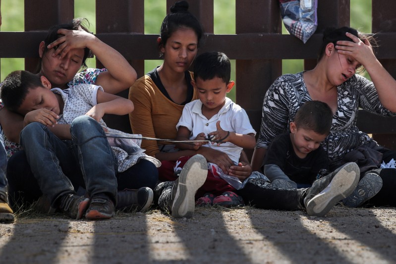 Immigrant women hold their children along the border wall as they await apprehension after illegally crossing into the U.S. from Mexico