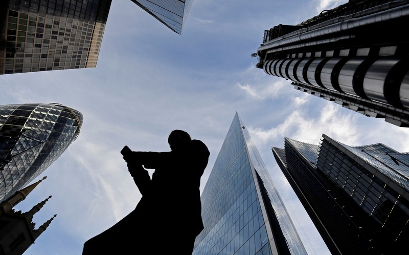 A worker looks at their phone as they walk past The Gherkin, Lloyds, and other office buildings in the City of London