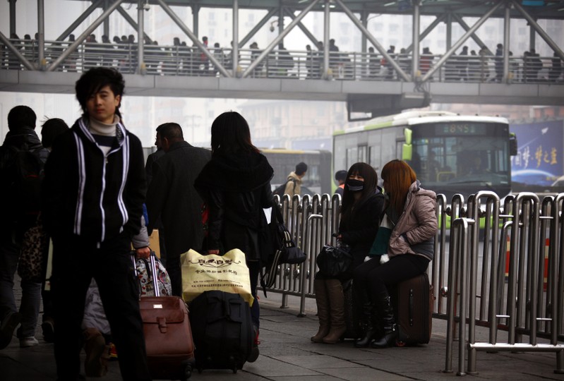 Pedestrians walk past two girls, one wearing a face mask, sitting on their luggage outside the Beijing west railway station