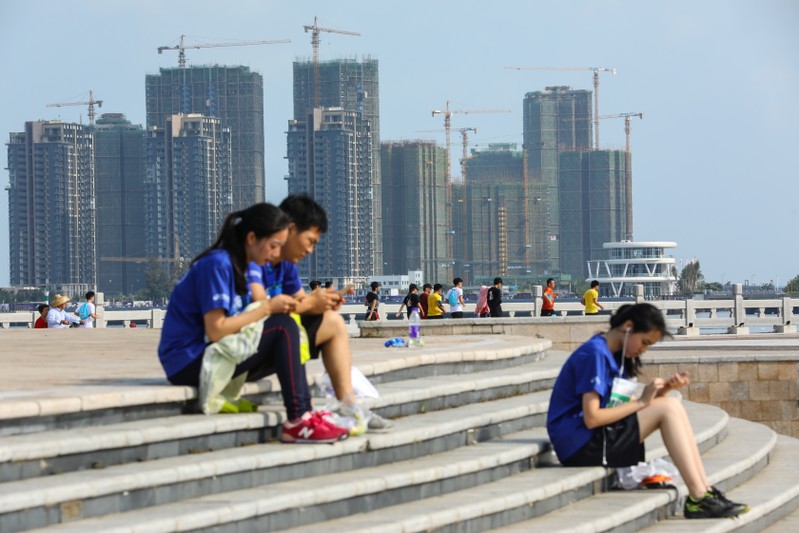 People sit on steps against a backdrop of residential buildings under construction, in Haikou