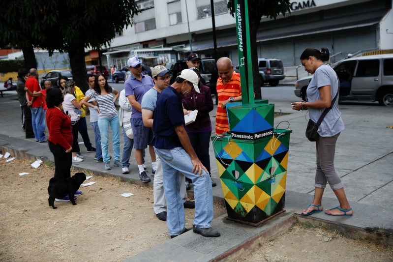 People line up as others charge their phones with a solar panel at a public square in Caracas