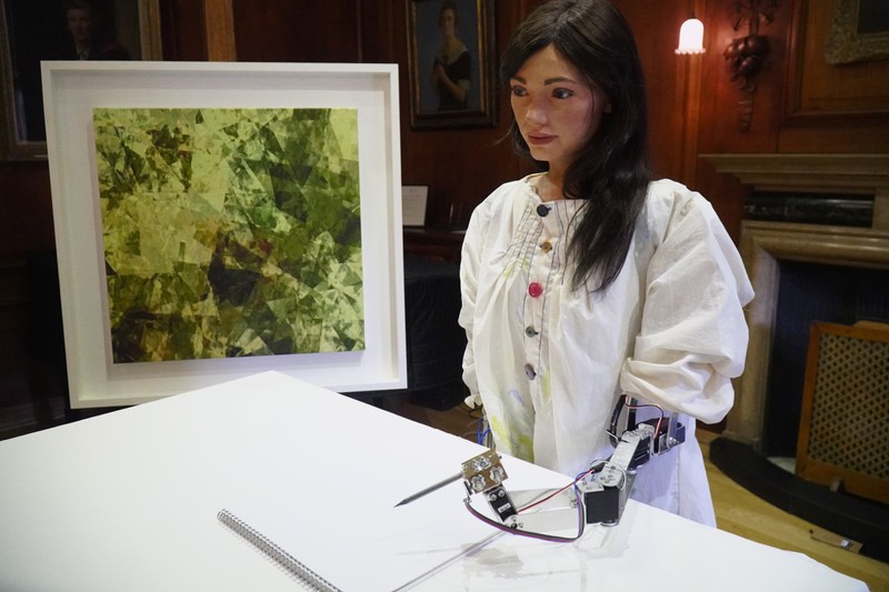 Robot artist 'Ai-Da' sketches using a pencil attached to her robotic arm, while standing next to a painting based on her computer vision data when run through algorithms developed by computer scientists in Oxford