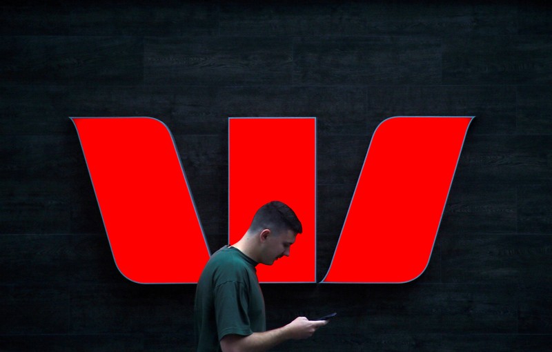 FILE PHOTO: A pedestrian looks at his phone as he walks past a logo for Australia's Westpac Banking Corp located outside a branch in central Sydney