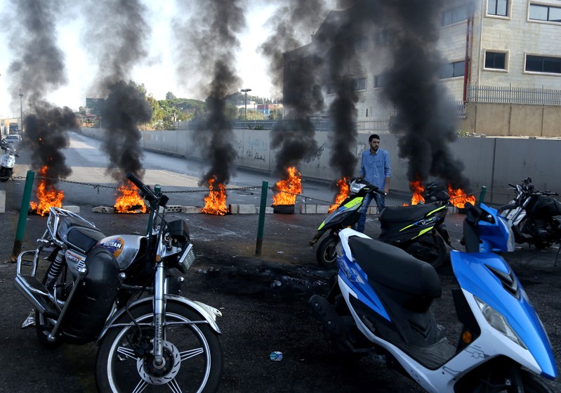 A demonstrator walks past a closed road by burning tires during ongoing anti-government protests, in Khaldeh