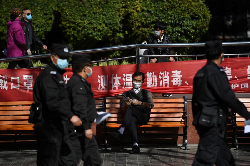 Police officers walk past a man holding his mobile phone while wearing face mask and plastic gloves, on a street in Kunming