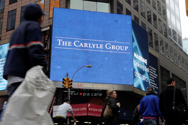 Passersby walk in front of video monitors announcing the Carlyle Group's listing on the NASDAQ market site in New York's Times Square after the opening bell for trading