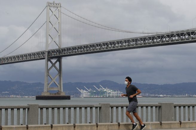 A man wears a mask to protect himself from the coronavirus while running in front of the San Francisco-Oakland Bay Bridge. | AP Photo