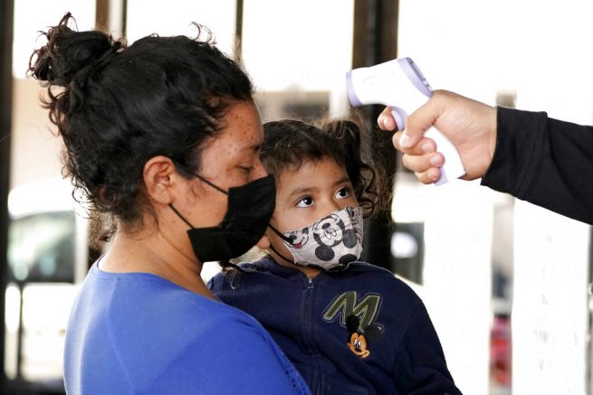 A migrant's temperature is taken at the entrance of a respite center as a child looks on after they were taken into custody and released from U.S. Customs and Border Protection while trying to sneak into the U.S., Friday, March 19, 2021, in McAllen, Texas. A surge of migrants on the Southwest border has the Biden administration on the defensive. The head of Homeland Security acknowledged the severity of the problem Tuesday but insisted it's under control and said he won't revive a Trump-era practice of immediately expelling teens and children. (AP Photo/Julio Cortez)