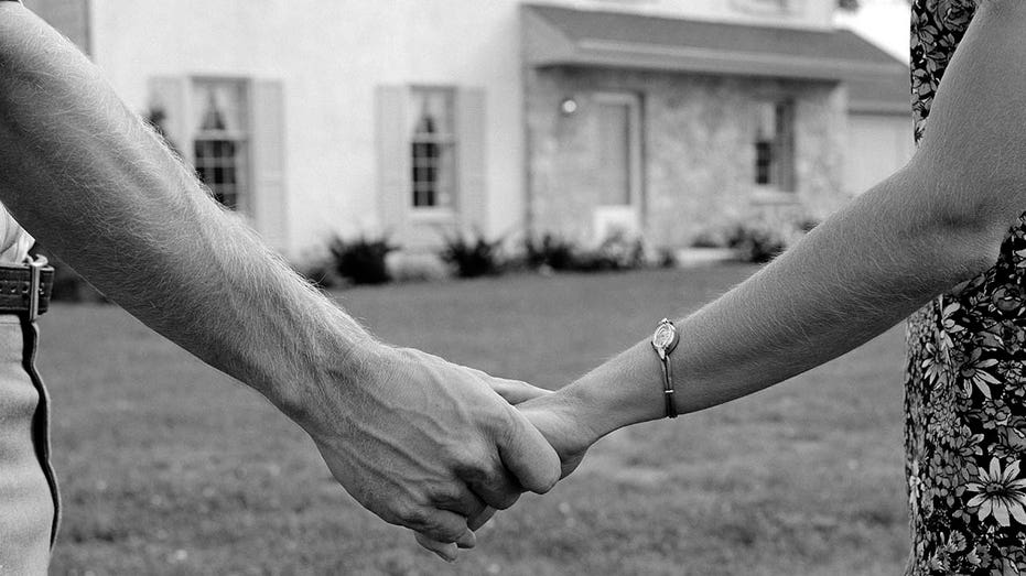 1970s close-up of a couple holding hands with their new home in the background.