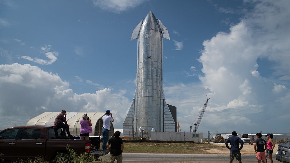 A silver prototype of SpaceX's Starship spacecraft in front of a blue sky as people in the foreground look on