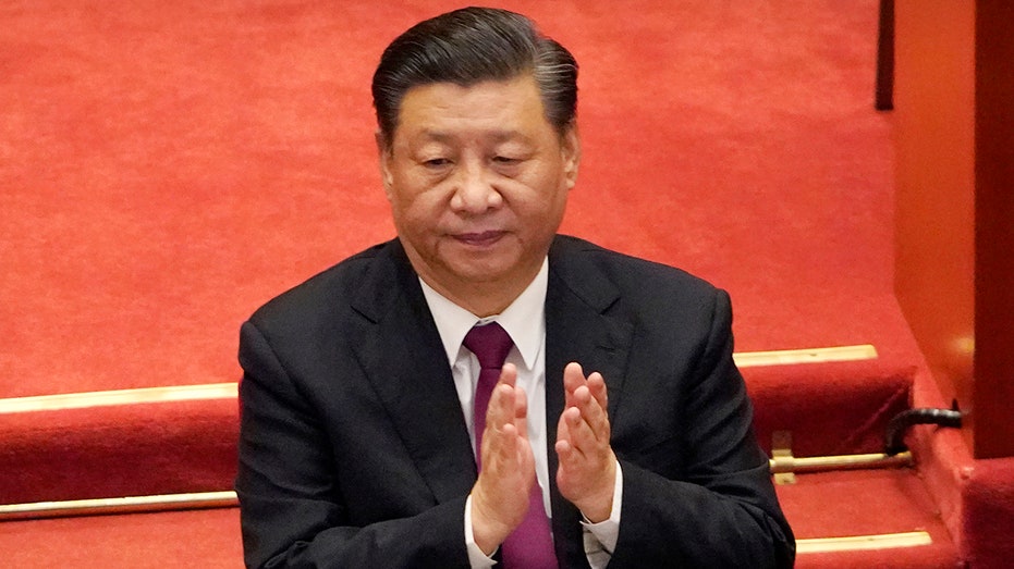 Chinese President Xi Jinping clapping wearing a suit and burgundy tie, sitting in front of a red background