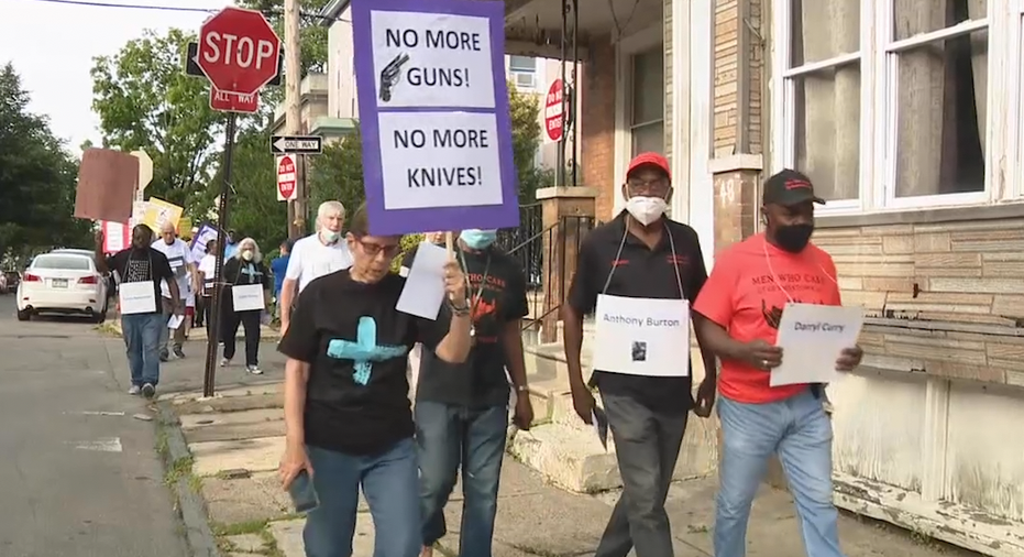 Woman walking with other protesters holding a sign that says 'NO MORE GUNS, NO MORE KNIVES'