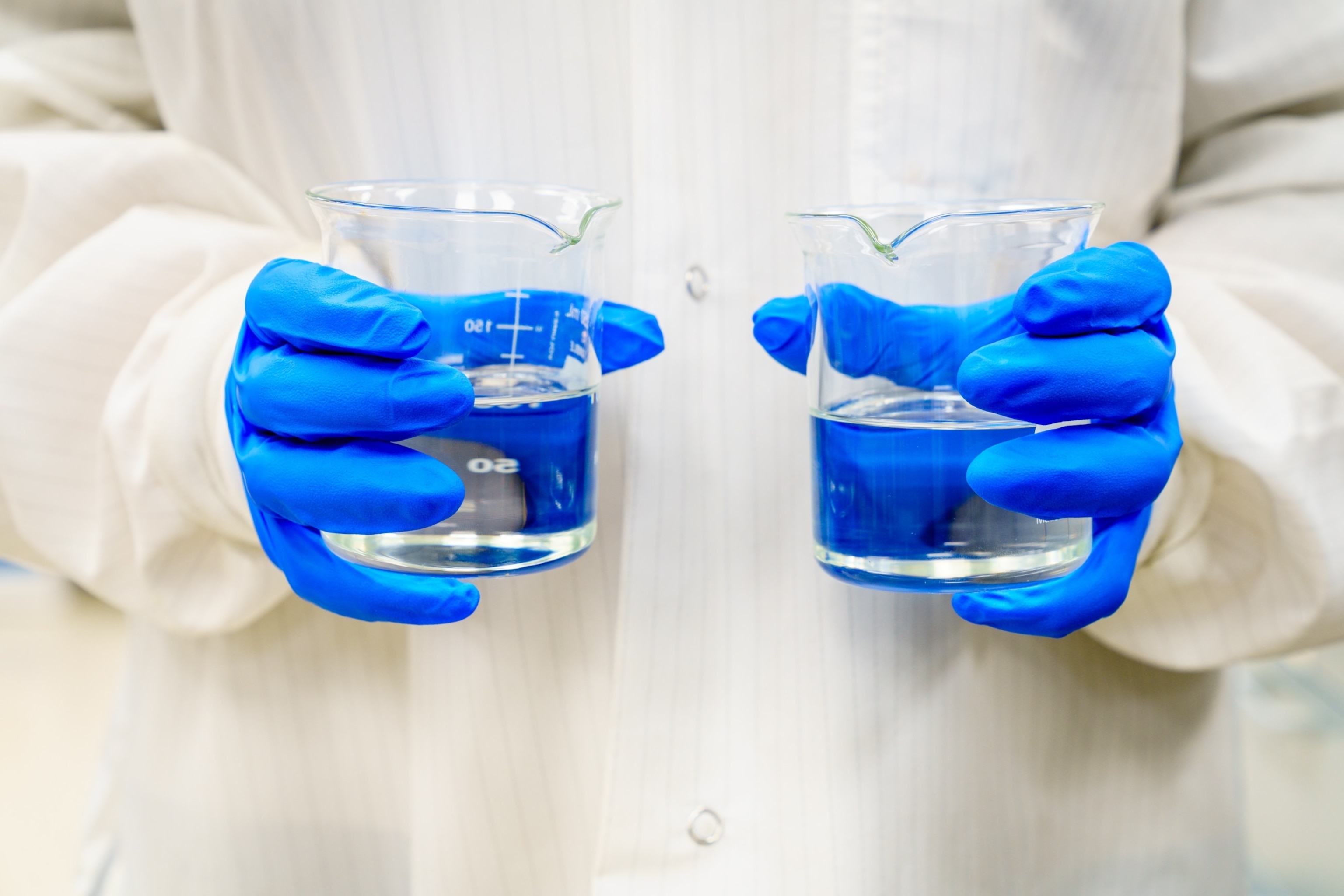 PHOTO: A person in a lab coat holding two beakers with clear liquids.