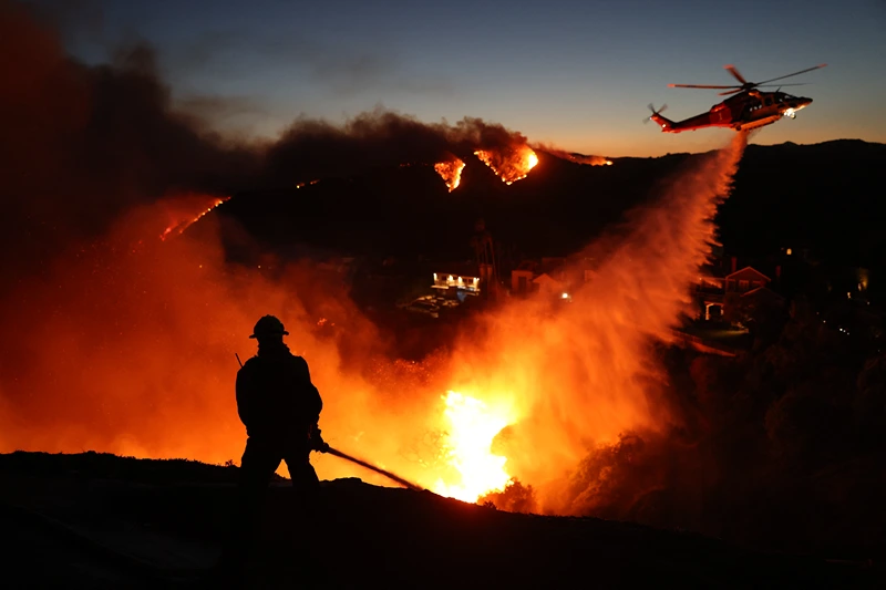 TOPSHOT - Fire personnel respond to homes destroyed while a helicopter drops water as the Palisades Fire grows in Pacific Palisades, California on January 7, 2025. A fast-moving wildfire in a Los Angeles suburb burned buildings and sparked panic, with thousands ordered to evacuate January 7, 2025 as "life threatening" winds whipped the region. Frightened residents abandoned their cars on one of the only roads in and out of the upscale Pacific Palisades area, fleeing on foot from the 770-acre (310-hectare) blaze engulfing an area crammed with multi-million dollar homes in the Santa Monica Mountains. (Photo by David Swanson / AFP) (Photo by DAVID SWANSON/AFP via Getty Images)